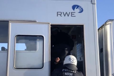 Ein behelmter Polizist schiebt einen Gefesselten die Treppe eines silbernen Gefangenentransporters mit RWE-Logo hoch