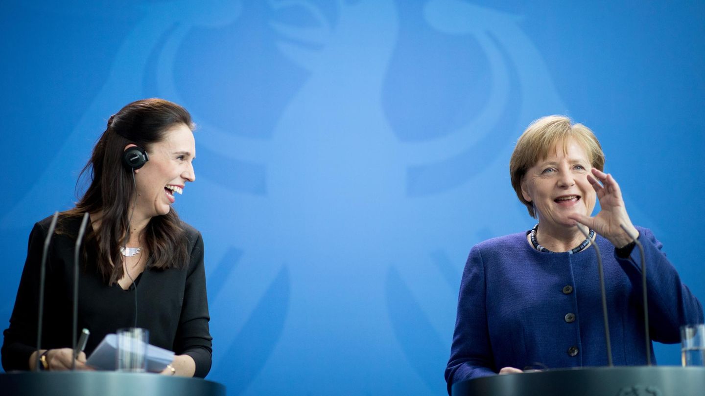 Jacinda Ardern und Angela Merkel stehen an zwei Rednerpulten und lachen vor einer blauen Wand mit hellblauem Bundesadler