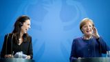 Jacinda Ardern und Angela Merkel stehen an zwei Rednerpulten und lachen vor einer blauen Wand mit hellblauem Bundesadler