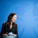 Jacinda Ardern und Angela Merkel stehen an zwei Rednerpulten und lachen vor einer blauen Wand mit hellblauem Bundesadler