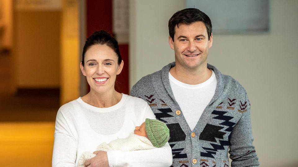 Jacinda Ardern steht mit einem Baby auf dem Arm vor einem Haus. Neben ihr steht ihr Partner Clarke Gayford