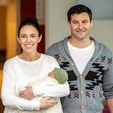 Jacinda Ardern steht mit einem Baby auf dem Arm vor einem Haus. Neben ihr steht ihr Partner Clarke Gayford