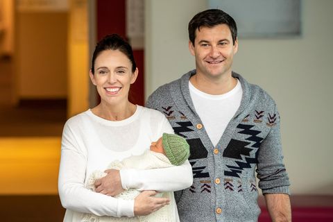 Jacinda Ardern steht mit einem Baby auf dem Arm vor einem Haus. Neben ihr steht ihr Partner Clarke Gayford