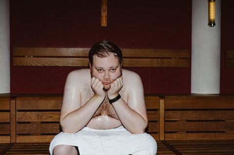 Ein Mann sitzt mit weißem Handtuch in der Sauna und schwitzt, den Kopf auf die Hände gestützt