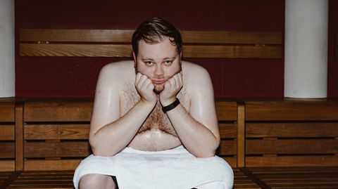 Ein Mann sitzt mit weißem Handtuch in der Sauna und schwitzt, den Kopf auf die Hände gestützt