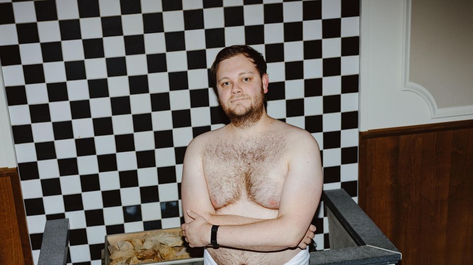 Reporter Rune Weichert in der Sauna Ein Mann mit verschränkten Armen und in einem weißen Handtuch steht vor einem Saunaofen, im Hintergrund schwarz-weiße Kacheln