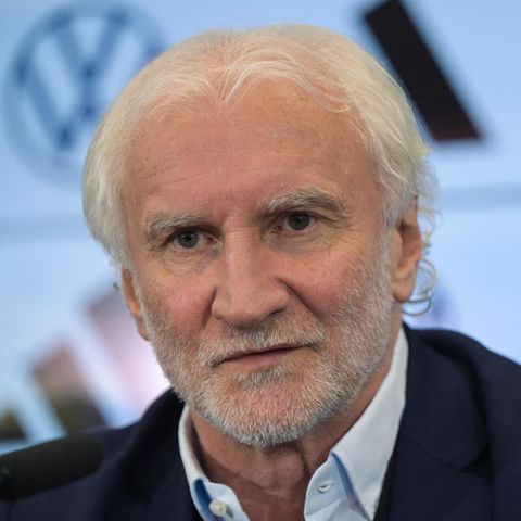 Rudi Völler, ein weißer Mann mit weißen Locken und Drei-Tage-Bart, sitzt in Hemd und Jackett auf einem Podium und spricht