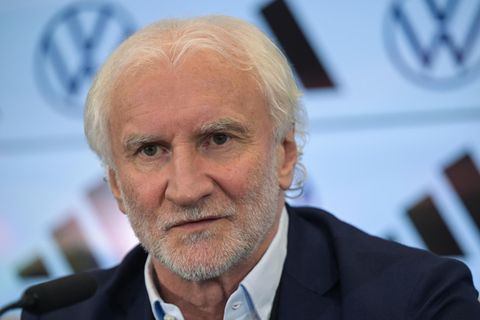 Rudi Völler, ein weißer Mann mit weißen Locken und Drei-Tage-Bart, sitzt in Hemd und Jackett auf einem Podium und spricht