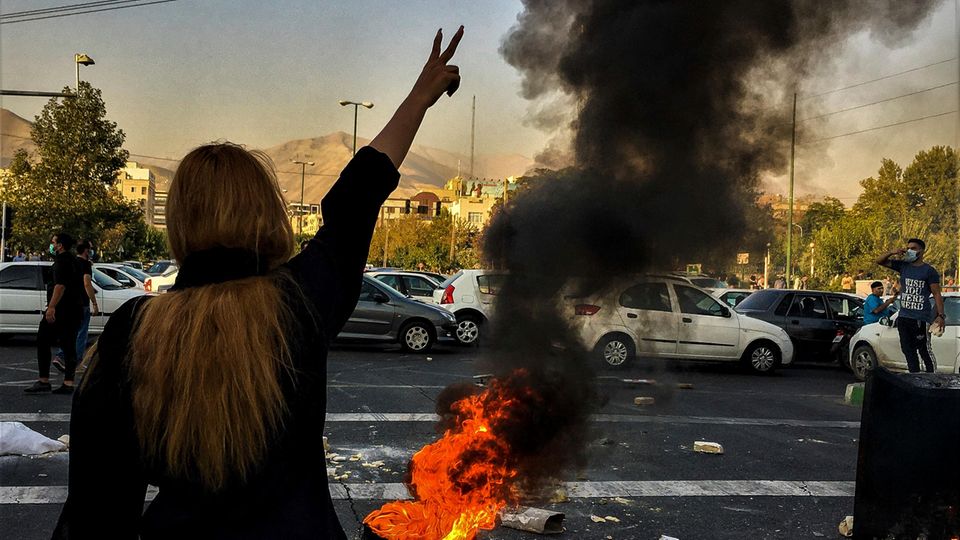 Im Iran steht eine Frau ohne Kopftuch vor einem brennenden Autoreifen auf einer Straße und macht mit rechts ein Victory-Zeichen