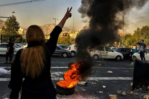 Im Iran steht eine Frau ohne Kopftuch vor einem brennenden Autoreifen auf einer Straße und macht mit rechts ein Victory-Zeichen