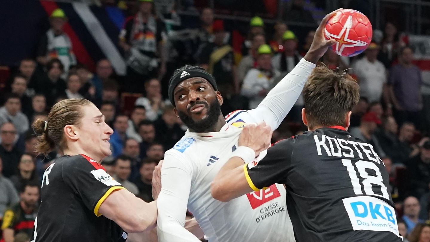 Ein schwarzer Handballer in weißem Trikot springt mit rotem Ball zwischen zwei weißen Handballern in schwarz hoch