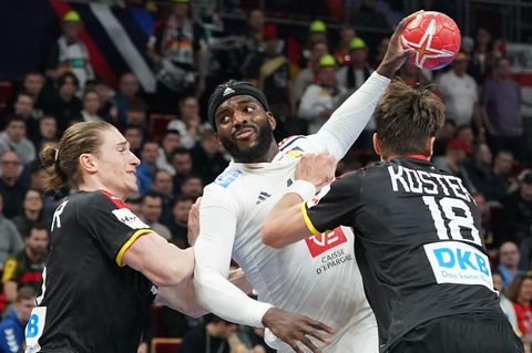 Ein schwarzer Handballer in weißem Trikot springt mit rotem Ball zwischen zwei weißen Handballern in schwarz hoch