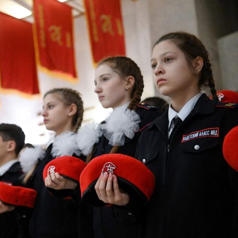 Mädchen stehen in dunkelblauen Uniformen in Reih und Glied und halten ein rotes Barett vor sich