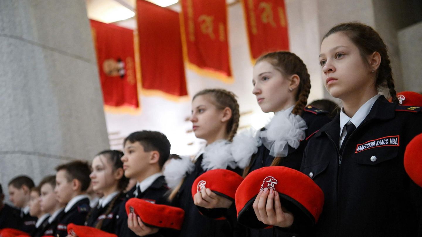 Mädchen stehen in dunkelblauen Uniformen in Reih und Glied und halten ein rotes Barett vor sich