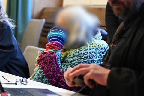 Eine ältere Frau mit weißen, schulterlangen Haaren sitzt in einem bunt gemusterten Strickpulli in einem Gerichtssaal