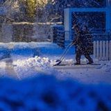Im bläulichen Licht des Morgens schiebt ein Mann in Winterkleidung Schnee von einem Fußweg, während weiter Schneeflocken fallen