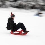 Ein Mann in schwarz fährt auf einem roten Schlitten einen Berg hinunter, schräg hinter ihm fahren ein Kind und ein Erwachsener