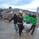Vier Männer tragen einen weißen Sack mit einem menschlichen Körper darin über eine schlammige Straße in Syrien