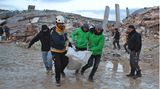 Vier Männer tragen einen weißen Sack mit einem menschlichen Körper darin über eine schlammige Straße in Syrien