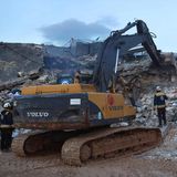 Ein Bagger trägt einen Trümmerhaufen ab, während drei Retter mit weißen Helmen genau hinschauen