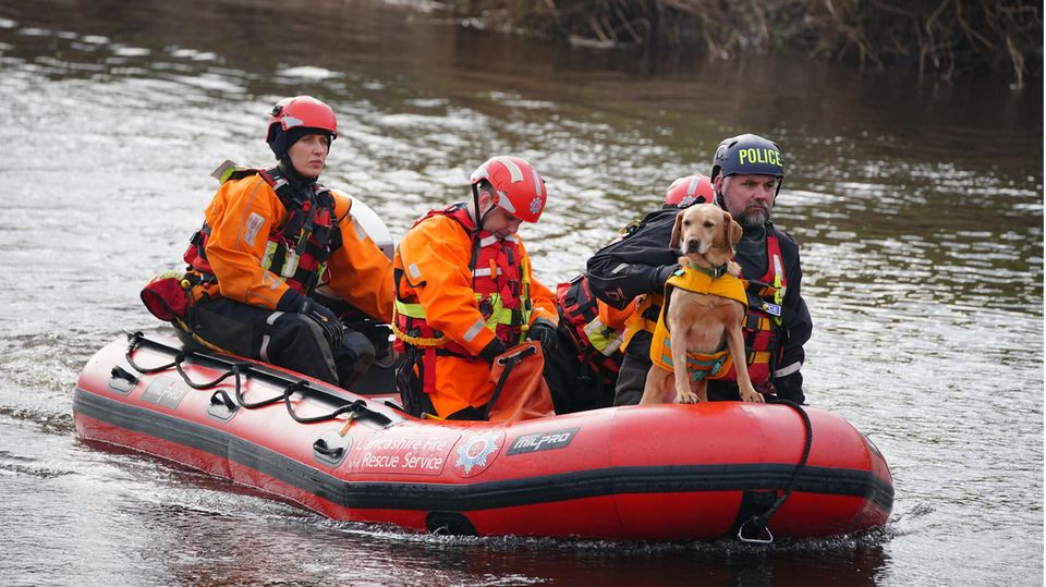 In einem roten Polizei-Schlauchboot auf dem Fluss Wyre sitzen vier Personen in oranger Kleidung. Ein blonder Hund sitzt im Bug