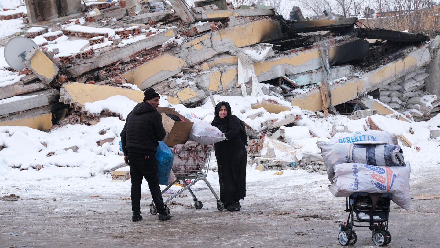 Ein Mann und eine Frau in Winterkleidung schieben einen vollen Einkaufswagen an einem eingestürzten Gebäude in der Türkei vorbei