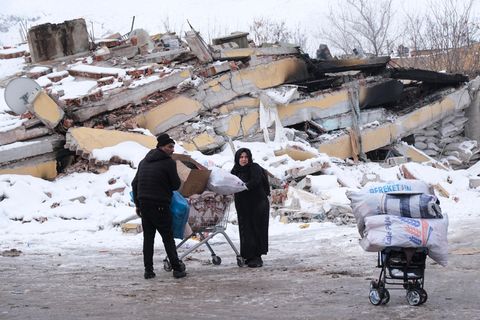 Ein Mann und eine Frau in Winterkleidung schieben einen vollen Einkaufswagen an einem eingestürzten Gebäude in der Türkei vorbei
