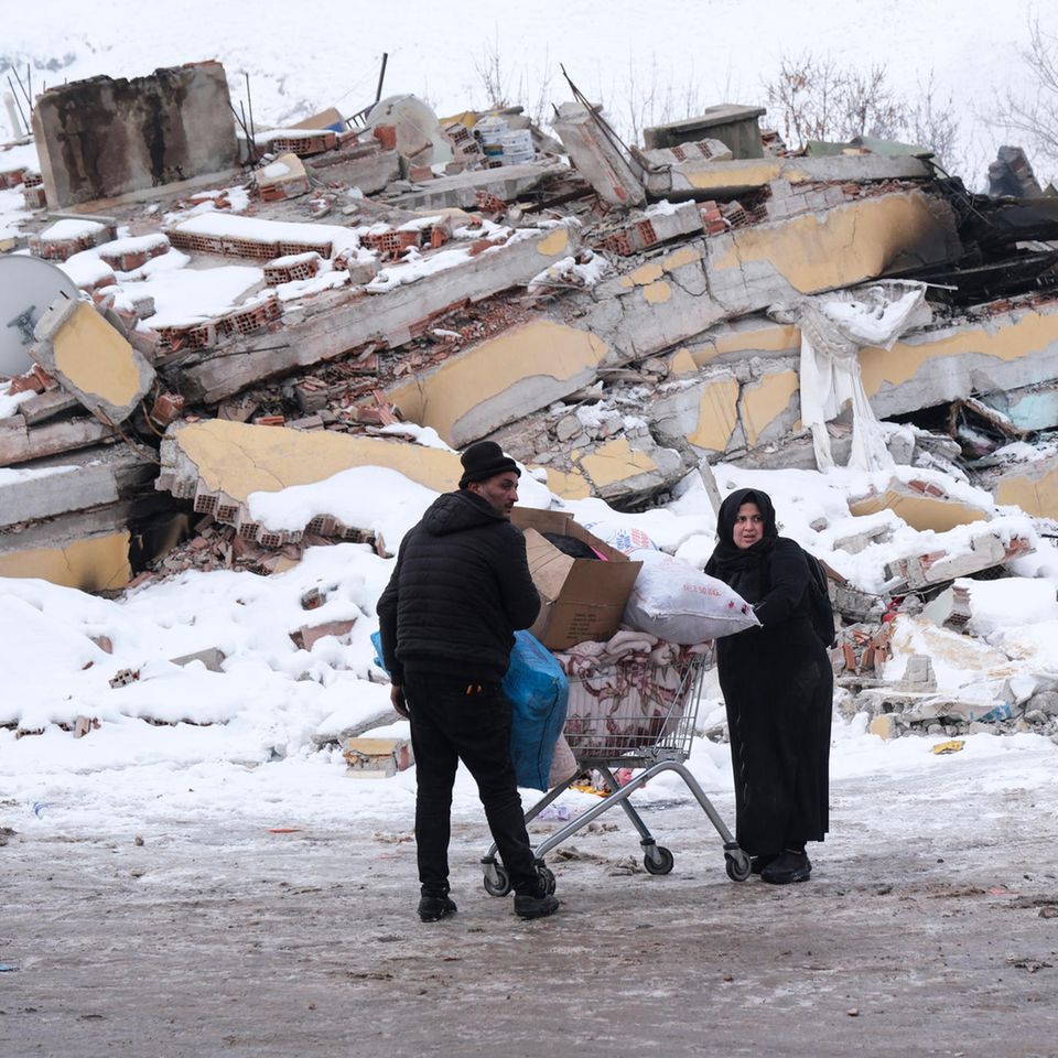 Ein Mann und eine Frau in Winterkleidung schieben einen vollen Einkaufswagen an einem eingestürzten Gebäude in der Türkei vorbei