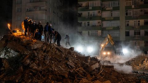 Mehrere Menschen stehen auf einem Trümmerberg, an dessen Fuß ein gelber Bagger mit eingeschalteten Scheinwerfern arbeitet