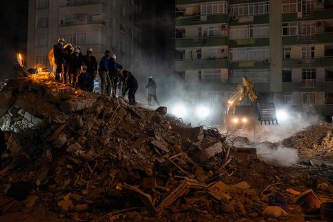 Mehrere Menschen stehen auf einem Trümmerberg, an dessen Fuß ein gelber Bagger mit eingeschalteten Scheinwerfern arbeitet