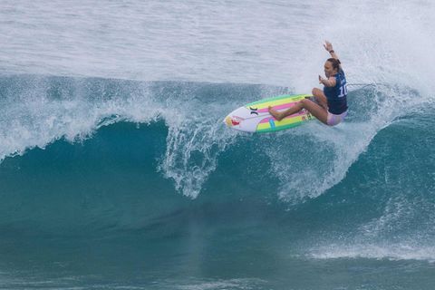 Eine Frau auf einem bunt gestreiften Surfbrett reitet mit ausgebreiteten Armen eine brechende Welle