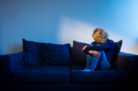 Eine Frau sitzt zusammengekauert mit angezogenen Beinen und hängendem Kopf auf einem Sofa. Immer mehr Menschen gehen in Reha.