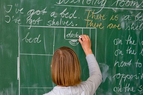 Eine Lehrerin mit schulterlangen blonden Haaren schreibt mit Kreide englische Wörter an eine grüne Tafel