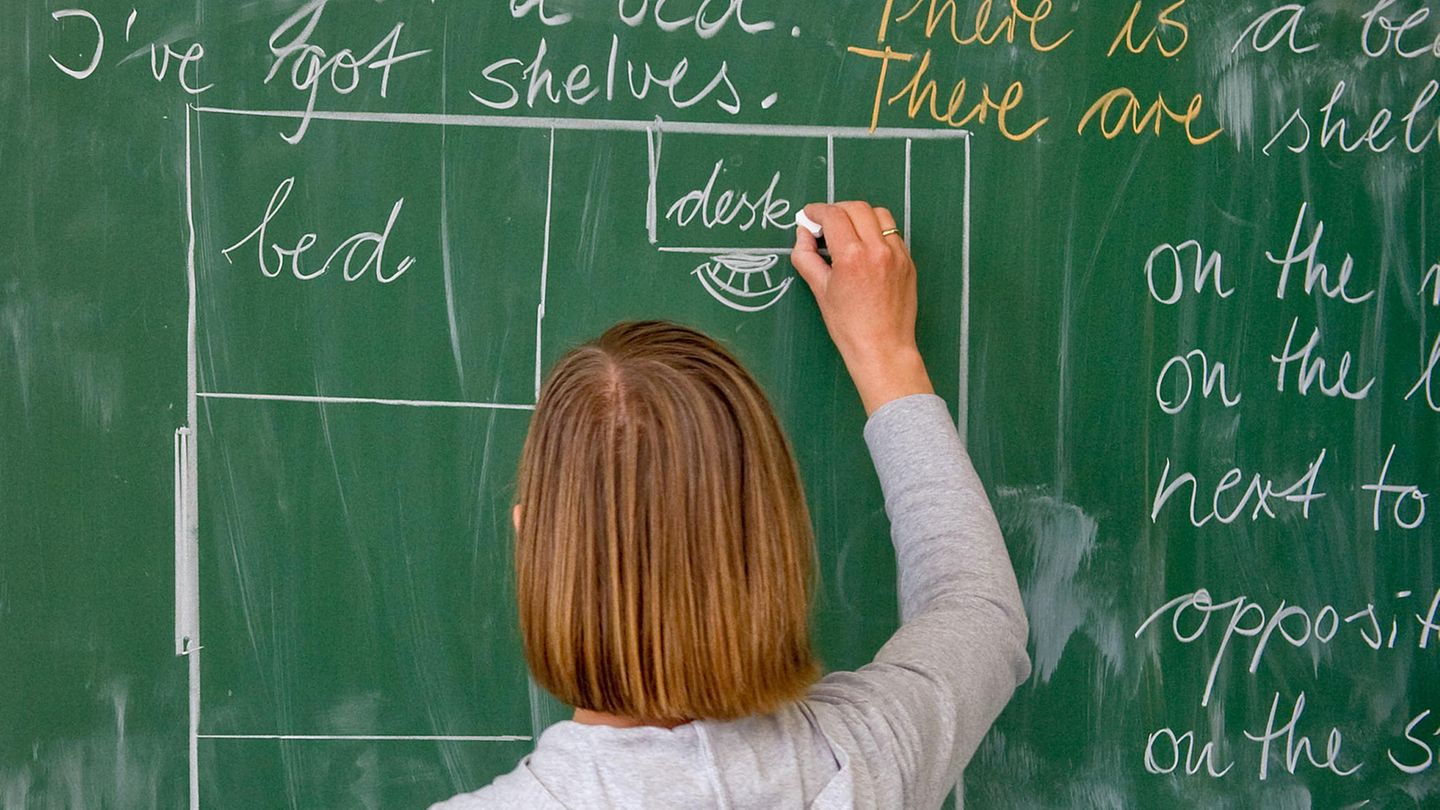 Eine Lehrerin mit schulterlangen blonden Haaren schreibt mit Kreide englische Wörter an eine grüne Tafel