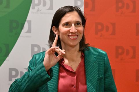 Elly Schlein, eine weiße Frau mit dunkelbraunen, schulterlangen Haaren und grünem Blazer, hebt lächelnd ihren Zeigefinger