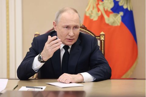 Russland: Putin sitzt an einem Tisch und hebt die Hand