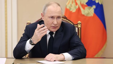 Russland: Putin sitzt an einem Tisch und hebt die Hand