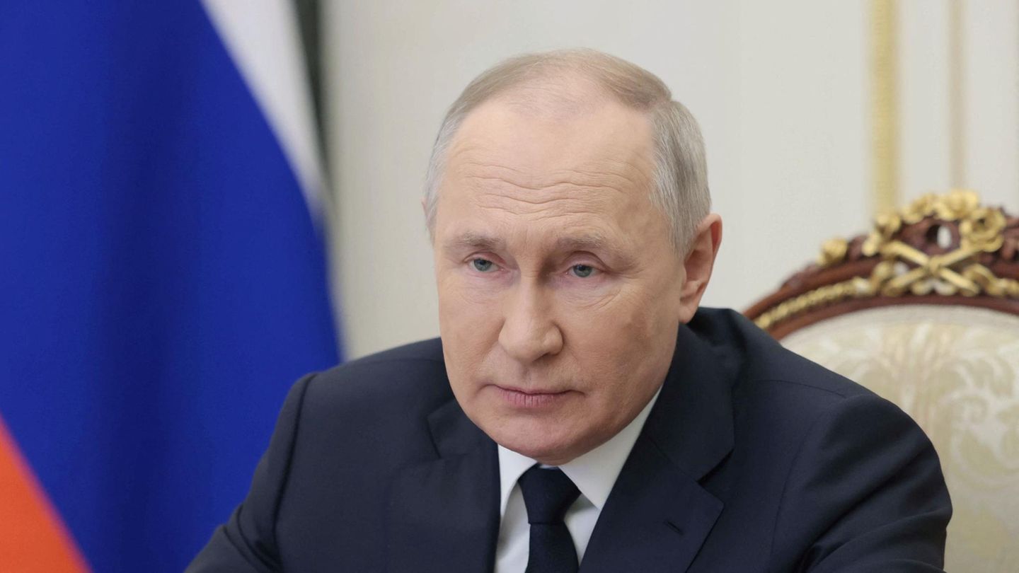 Wladimir Putin Wladimir Putin sitzt in Anzug und Krawatte auf einem antiken Stuhl vor einer russischen Flagge
