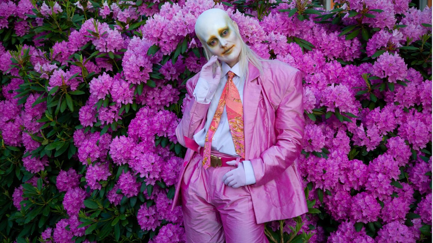 Sängerin Karin Dreijer von Fever Ray in einem pinken Anzug, mit weißer Schminke im Gesicht, vor einem Busch mit pinken Blüten