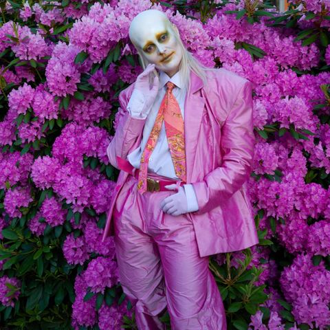 Sängerin Karin Dreijer von Fever Ray in einem pinken Anzug, mit weißer Schminke im Gesicht, vor einem Busch mit pinken Blüten