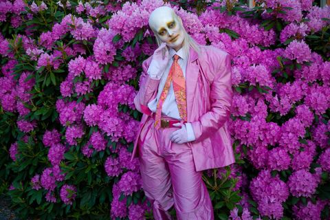 Sängerin Karin Dreijer von Fever Ray in einem pinken Anzug, mit weißer Schminke im Gesicht, vor einem Busch mit pinken Blüten