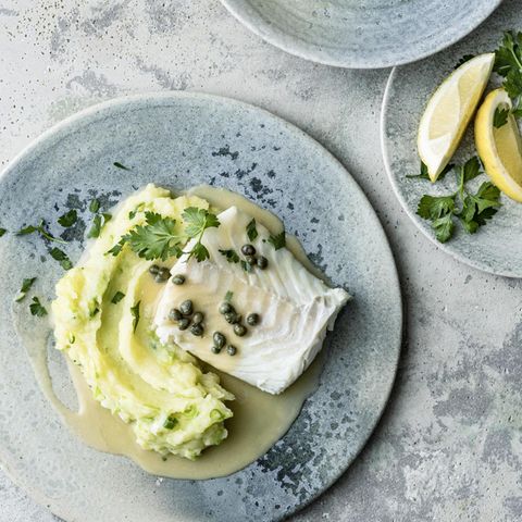 Auf blauen Tellern sind Kabeljau mit Kapernsauce und Kartoffelpüree, und Zitronenspalten zu sehen
