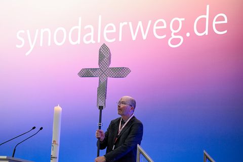 Vor dem Schriftzug "synodalerweg.de" wird auf der Synodalversammlung ein Kreuz auf dem Podium aufgestellt. 