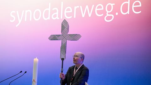 Vor dem Schriftzug "synodalerweg.de" wird auf der Synodalversammlung ein Kreuz auf dem Podium aufgestellt. 
