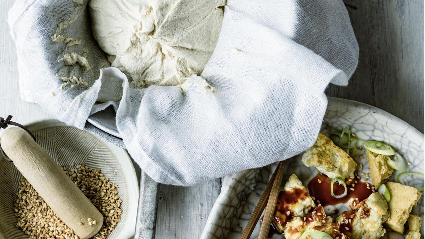 In drei hellen Schalen sind Sesamkörner, ein Mulltuch mit selbstgemachter Tofumasse, und fertiger Tofu mit einer Sauce