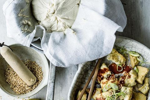 In drei hellen Schalen sind Sesamkörner, ein Mulltuch mit selbstgemachter Tofumasse, und fertiger Tofu mit einer Sauce