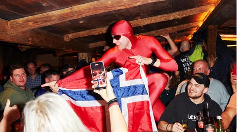 Ein Gast im roten Ganzkörperanzug hält in einer Bar in Ischgl eine Norwegen-Flagge hoch und wird fotografiert, drumherum Feiernd