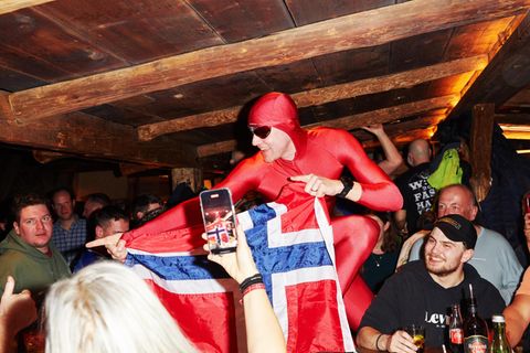 Ein Gast im roten Ganzkörperanzug hält in einer Bar in Ischgl eine Norwegen-Flagge hoch und wird fotografiert, drumherum Feiernd