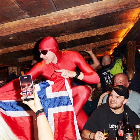 Ein Gast im roten Ganzkörperanzug hält in einer Bar in Ischgl eine Norwegen-Flagge hoch und wird fotografiert, drumherum Feiernd