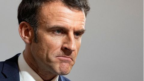 Emmanuel Macron, ein weißer Mann mit braunem Seitenscheitel, trägt blauen Anzug und Krawatte und schaut skeptisch